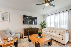 Ein modernes Wohnzimmer mit Sofa, Couchtisch und Sessel, einem Deckenventilator und einem großen Fenster mit Vorhängen.