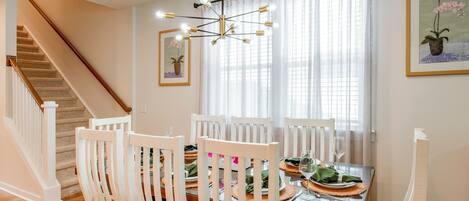 A dining area with a wooden staircase, white chairs, a glass table, and a chandelier.