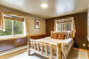 Ein Schlafzimmer mit einem hölzernen Bett, zwei mit Vorhängen versehenen Fenstern, einem gerahmten Bild an der Wand und einer Deckenleuchte.
