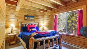 A wooden cabin room with a bed, a window with curtains, a lamp, and a painting on the wall. - Pool Table & Peaceful Porch: Fairplay Retreat (Fairplay)