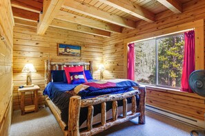 Ein Zimmer einer Holzhütte mit einem Bett, einem Fenster mit Vorhängen, einer Lampe und einem Bild an der Wand.