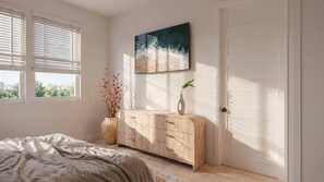 Une chambre à coucher avec un lit, une commode, un vase de fleurs, un tableau et une fenêtre avec des stores.
