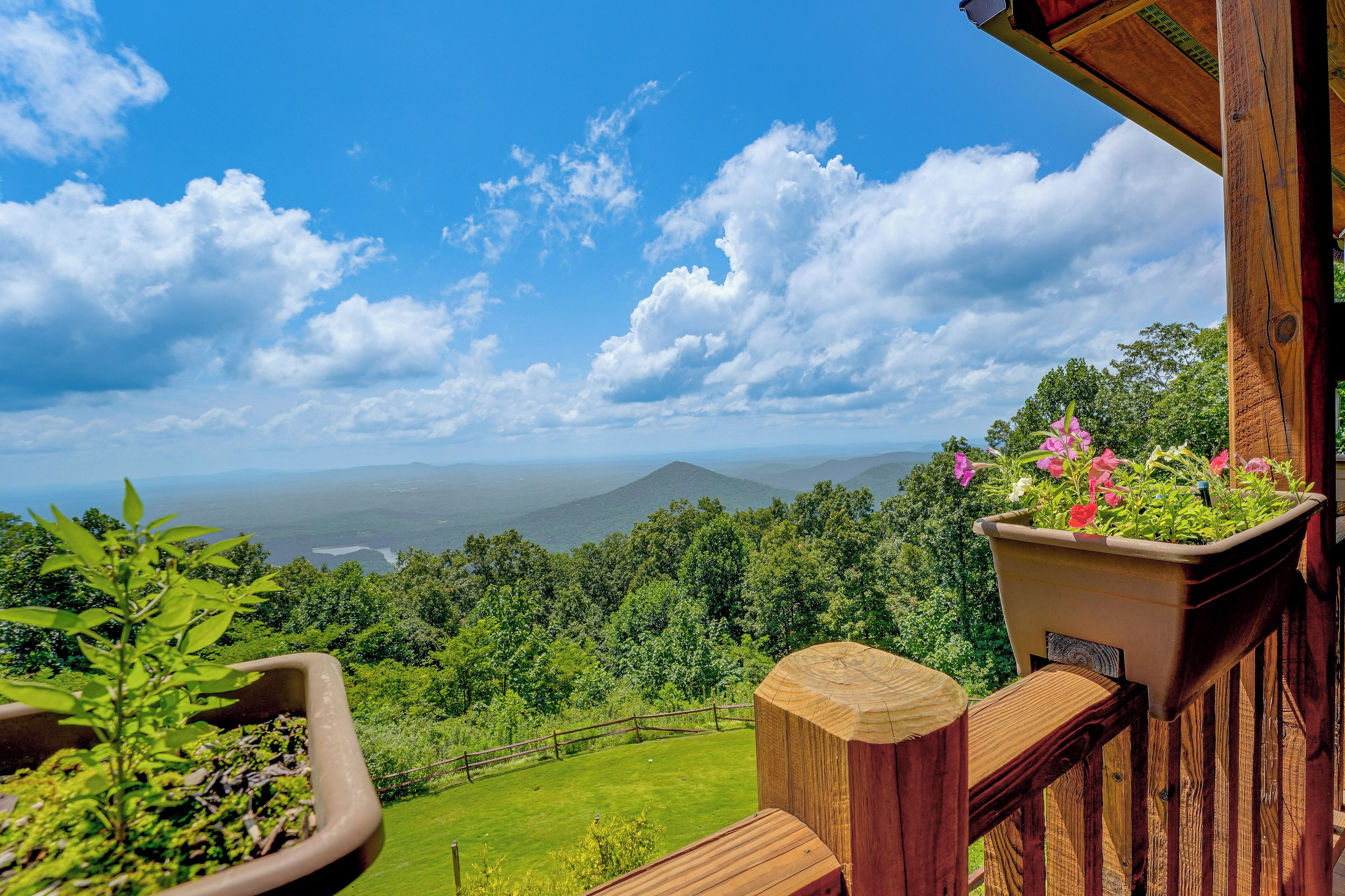 A balcony with a wooden railing overlooking a lush green landscape and distant mountains under a clear blue sky.