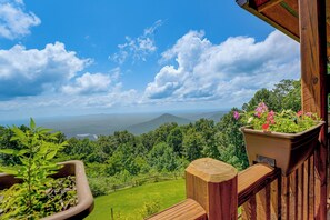 Un balcon doté d’une balustrade en bois, d’où l’on peut admirer un paysage verdoyant et luxuriant ainsi que des montagnes lointaines, le tout sous un ciel bleu dégagé.