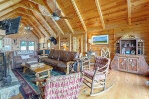 A rustic wooden interior with a high ceiling, a ceiling fan, a fireplace, a sofa, a rocking chair, and a cabinet with various items. - Mountain Top Lodge With Amazing Vista (Jasper)