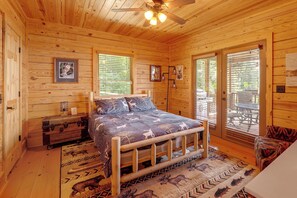 Ein Schlafzimmer einer Holzhütte mit einem Bett, einer Kommode, einem Nachttisch, einem Deckenventilator und einem Fenster mit Jalousien.