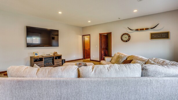 A living room with a large flat-screen TV mounted on the wall, a wooden entertainment center, a sofa with multiple cushions, and a clock on the wall. - Private Yard w/ Patio! Rapid City Escape (Rapid City)