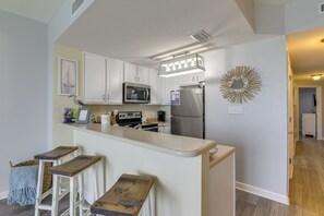 A kitchen with white cabinets, a stainless steel refrigerator, a microwave, and a stove. There are wooden stools at the counter. - Unwind Poolside, Walk to Sand! 18th-floor PCB Unit (Panama City Beach)