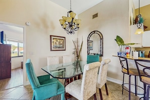 Un coin repas avec une table en verre, des chaises turquoise et blanches, un lustre et un comptoir de cuisine agrémenté d’une enseigne.