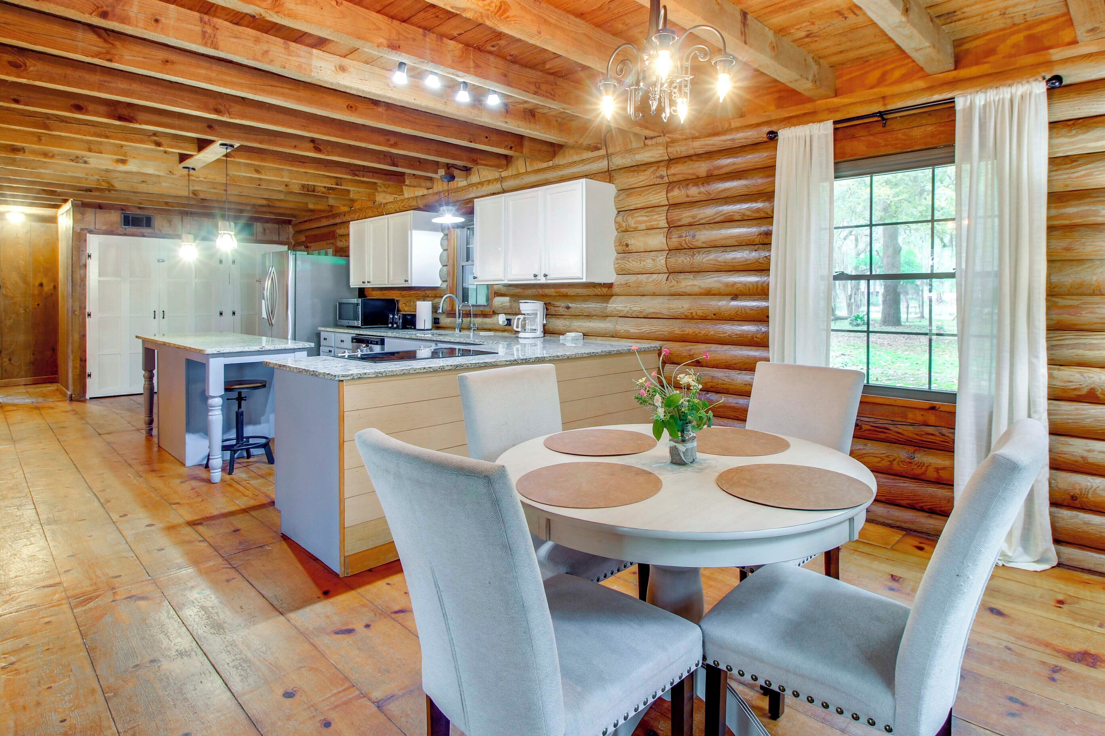 A log cabin interior with a central island kitchen, dining area with round tables, and a large window with curtains.