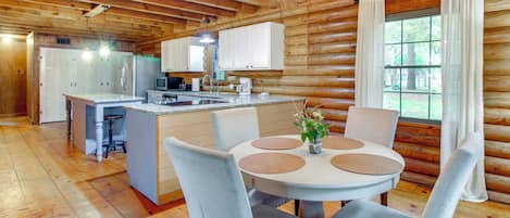 A log cabin interior with a central island kitchen, dining area with round tables, and a large window with curtains.