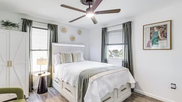 A bedroom with a bed, a bedside table, a chair, a ceiling fan, and a window with curtains.