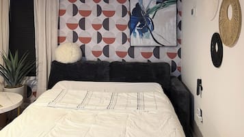 Et soveværelse med en seng, en rund hvid pude, en potteplante og en væg med et geometrisk mønster og et maleri.