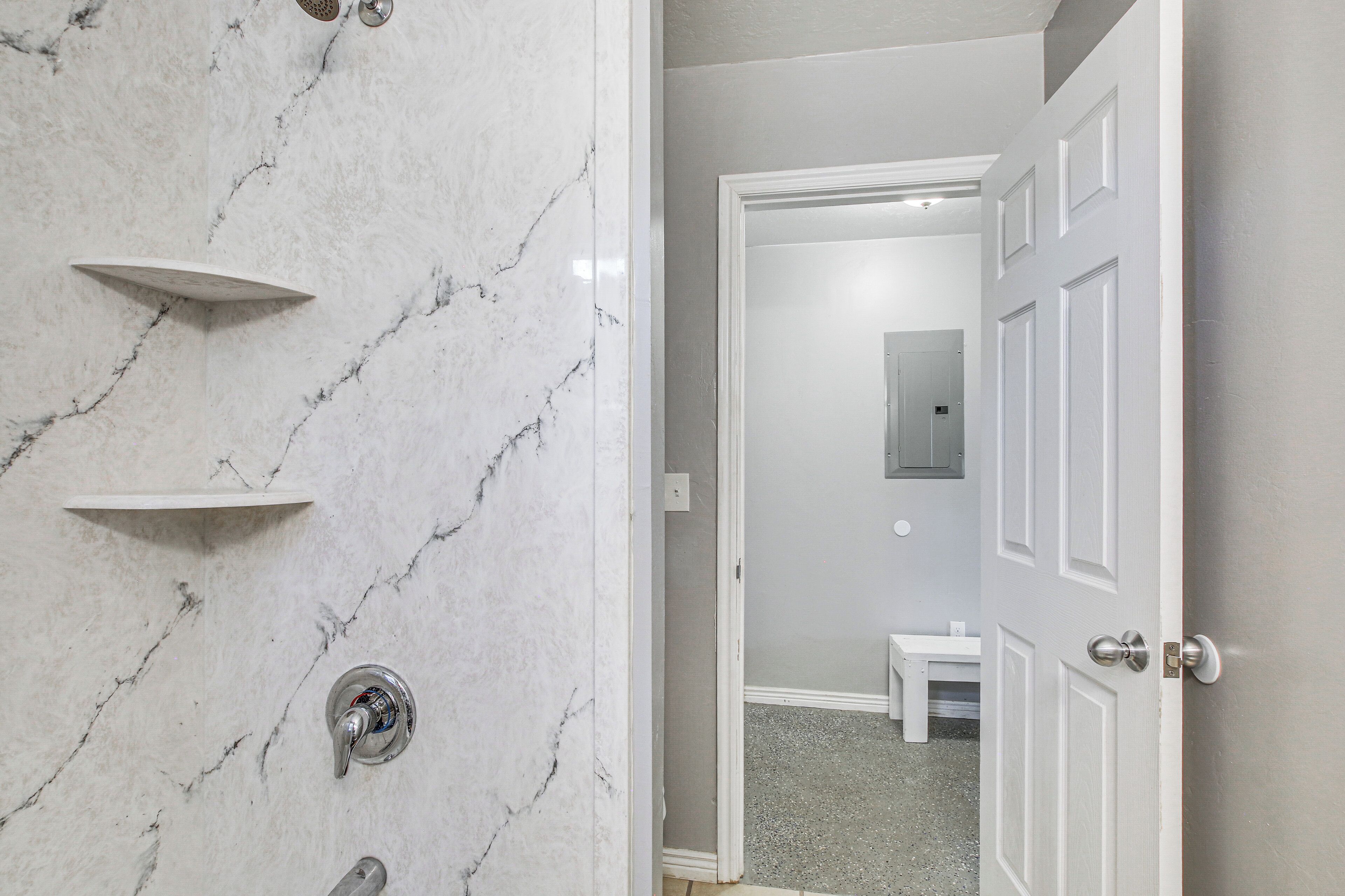 A bathroom with a marble wall, a shower with a control panel, and a door leading to a bedroom.