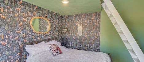 A bedroom with a bed, a mirror, a wall-mounted fan, and a ladder against a green wall.