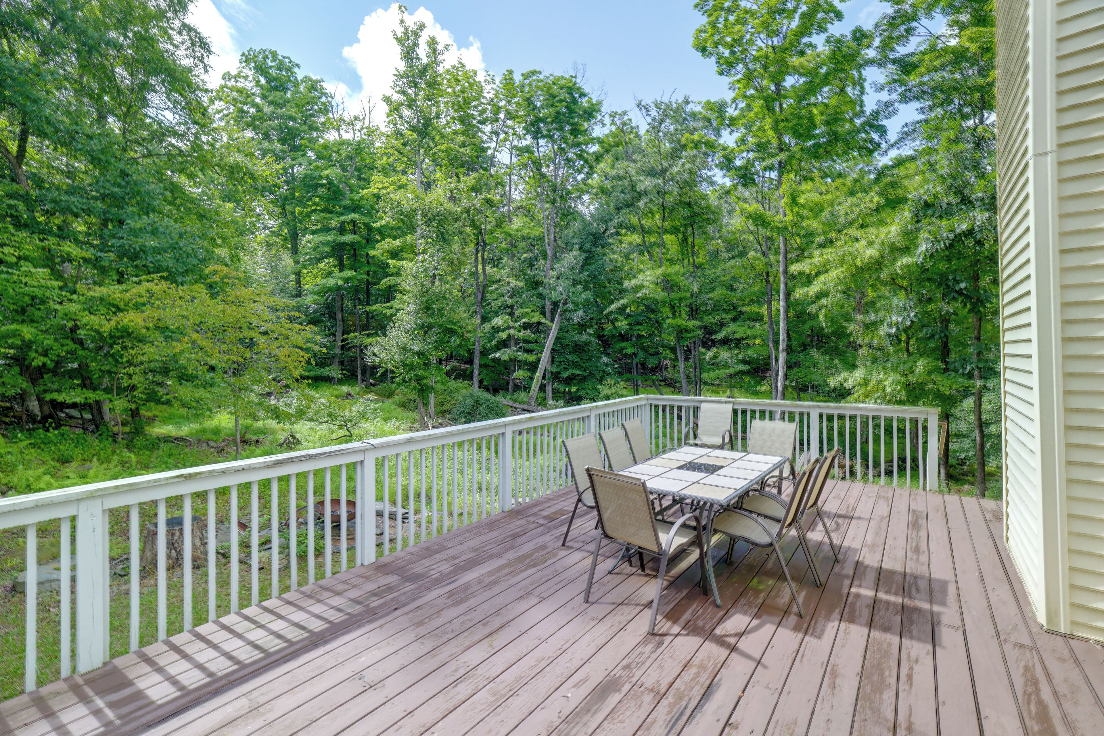 A wooden deck with a table and chairs, surrounded by a white railing, overlooking a forested area.