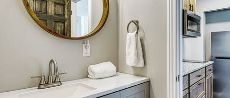 A bathroom with a sink, mirror, towel rack, and a door leading to a kitchen area.