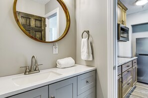 A bathroom with a sink, mirror, towel rack, and a door leading to a kitchen area.