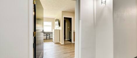 A hallway with a wooden floor, a white wall, and a door leading to a kitchen area.