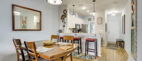 A dining area with a wooden table and chairs, a kitchen in the background, and a hallway leading to a door.