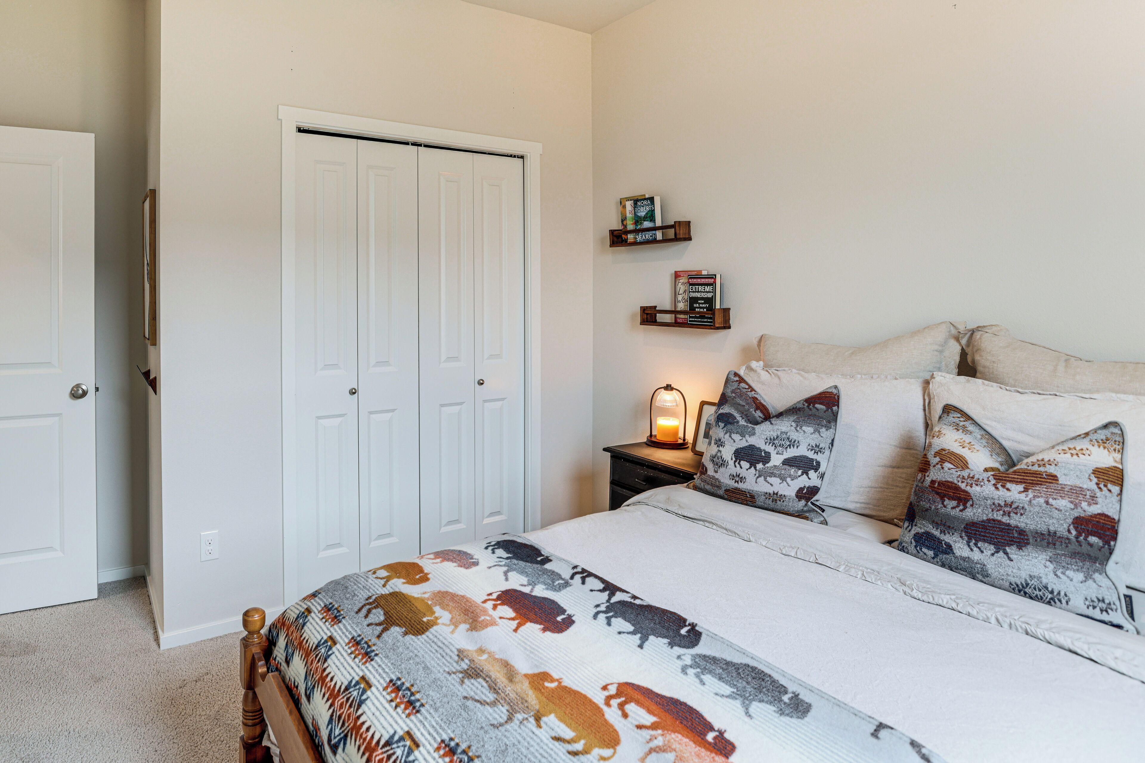 A bedroom with a bed, pillows, a nightstand, a wall-mounted shelf with books, and a closed door.