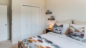 A bedroom with a bed, pillows, a nightstand, a wall-mounted shelf with books, and a closed door.