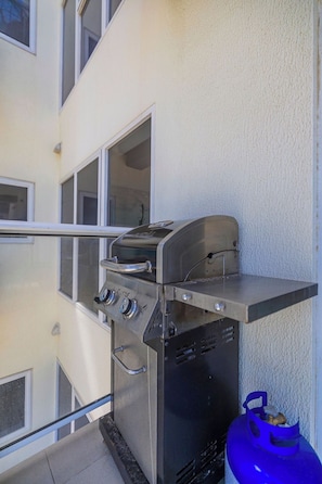A stainless steel outdoor grill with a gas tank attached, mounted on a balcony railing.