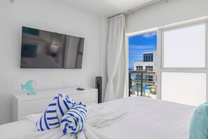A hotel room with a bed, a flat-screen TV mounted on the wall, a bedside table with a lamp, and a window with a view of buildings. - HH-2Bdr415 - Aruba's Amazing 2BDR Retreat. (Oranjestad)
