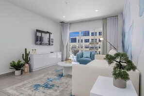 A modern living room with a sofa, a coffee table, a cactus plant, and a view of buildings through large windows. - HH-2Bdr415 - Aruba's Amazing 2BDR Retreat. (Oranjestad)