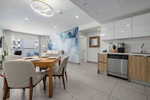A modern kitchen with a dishwasher, a dining area with a wooden table and chairs, and a living area with a sofa and a large abstract mural.