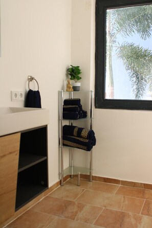 Ein Badezimmer mit einem weißen Waschbecken, einem Handtuchhalter, einer Glasablage mit Handtüchern, einer Topfpflanze und einem Fenster mit Blick auf Bäume.