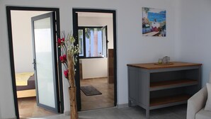 Uma porta aberta dá acesso a um quarto com cama e mesa de cabeceira, um vaso de madeira com flores e uma pintura de uma praia na parede.