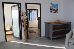 Eine geöffnete Tür, die in ein Schlafzimmer führt, mit einem Bett und einem Nachttisch, einer Holzvase mit Blumen und einem Bild einer Strandlandschaft an der Wand.