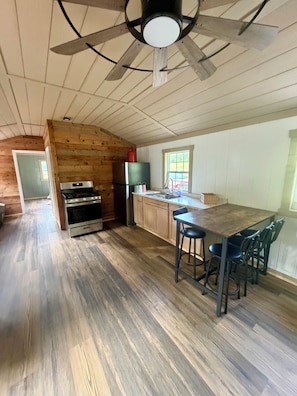 A kitchen with wooden floors, a central island with bar stools, a ceiling fan, and a window.