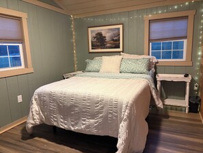 A bedroom with a bed, two bedside tables, a nightstand, a framed picture on the wall, and two windows with blinds.