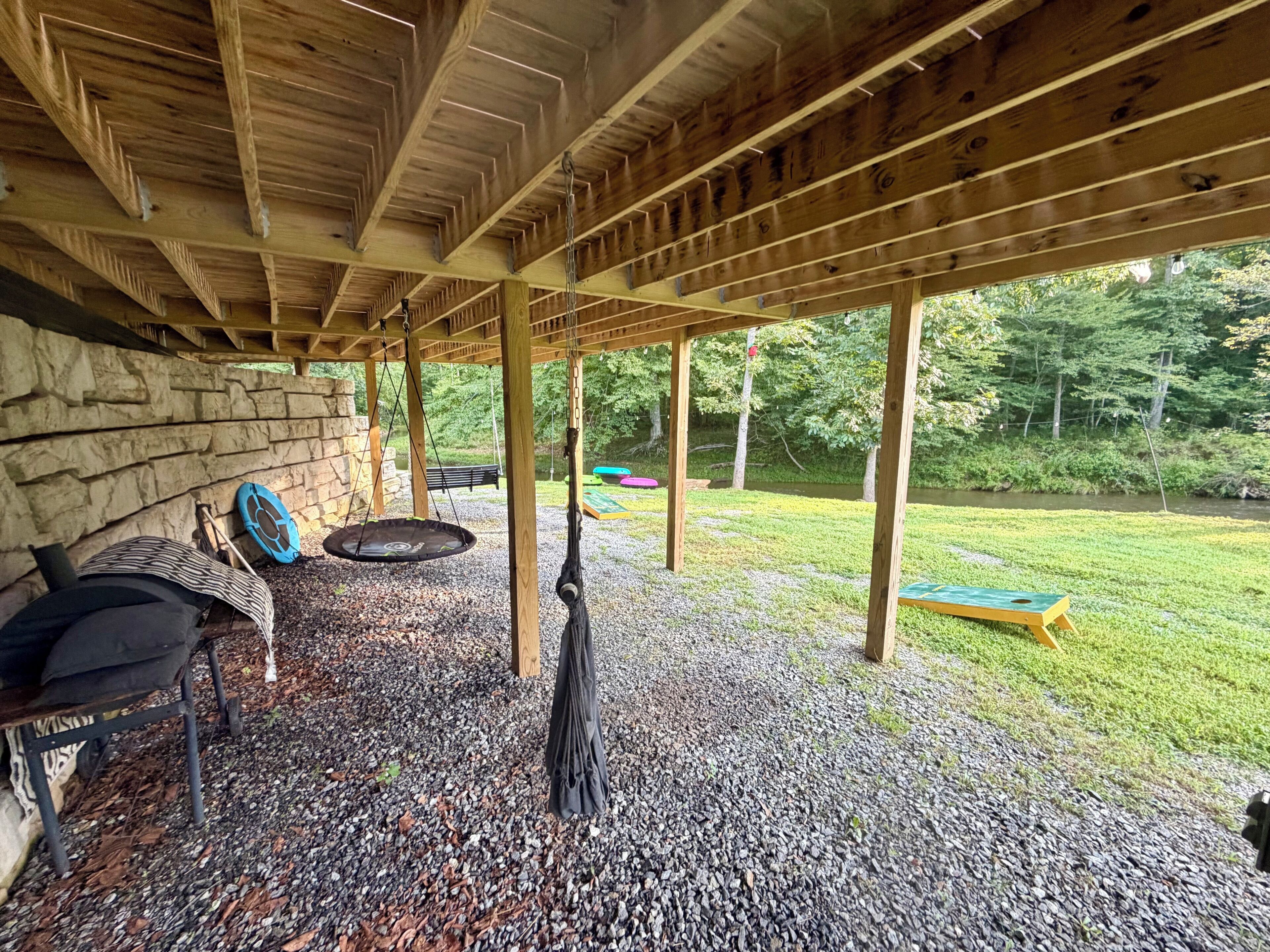 An outdoor covered patio with a stone wall, a hanging swing, a fire pit, and a grassy area with a bench.