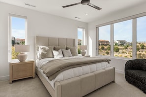 Ein Schlafzimmer mit Bett, Nachttischen, einer Lampe, einem Deckenventilator und großen Fenstern mit Blick auf eine Wohngegend.