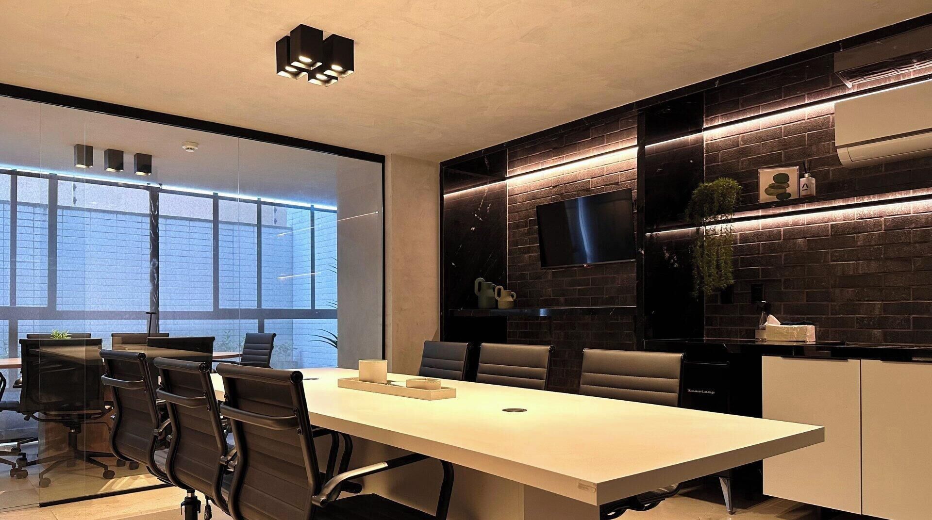 A modern conference room with a long table, multiple chairs, a flat-screen TV mounted on a brick wall, and a large window providing natural light.