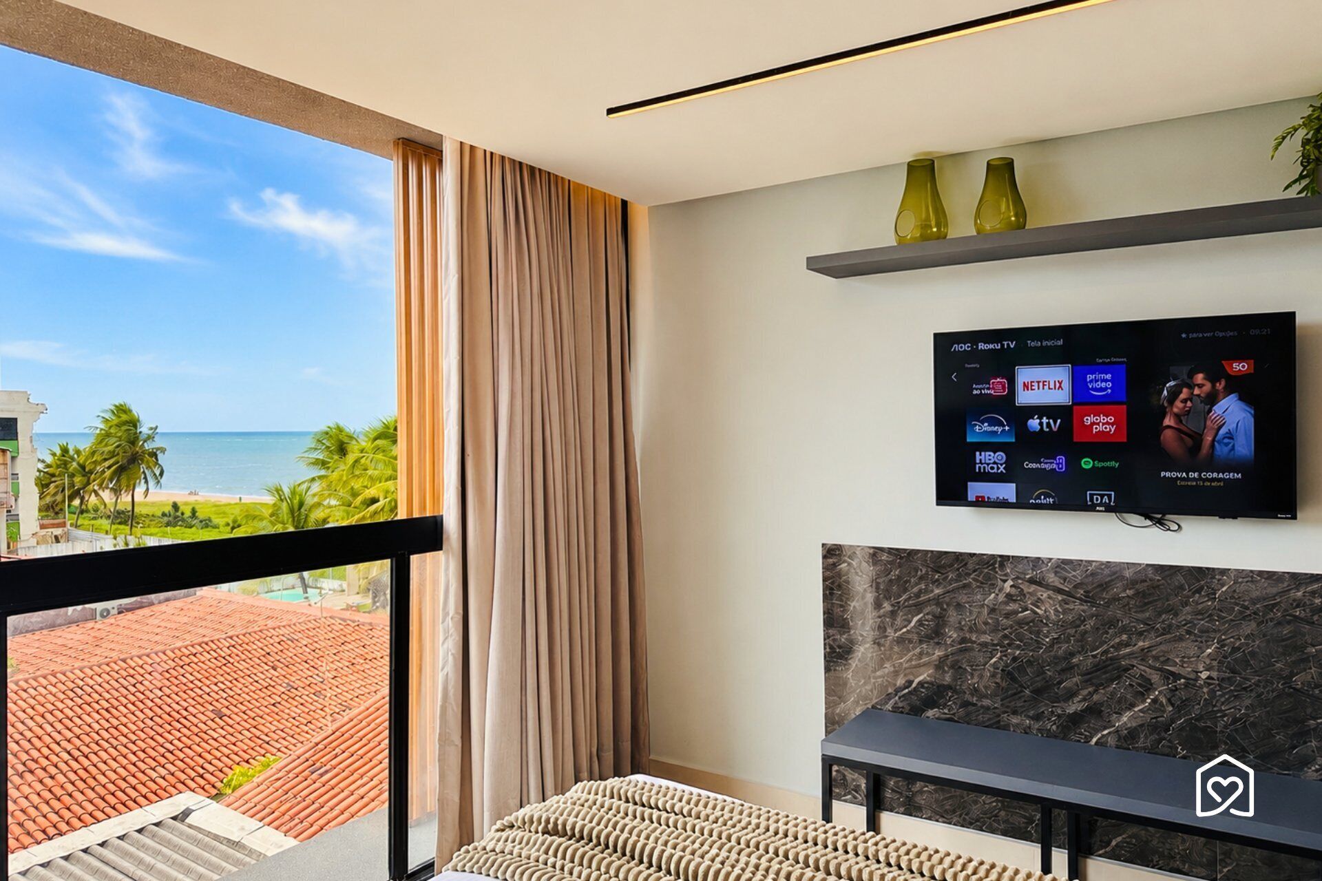 A hotel room with a large window, a flat-screen TV mounted on the wall, a small table, and a bench.