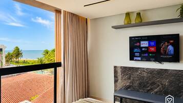 A hotel room with a large window, a flat-screen TV mounted on the wall, a small table, and a bench.