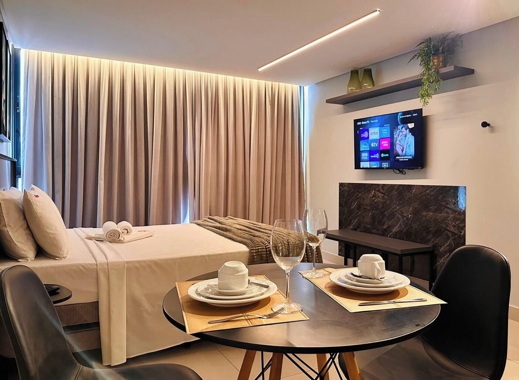 A hotel room with a bed, a dining table set for two, a TV mounted on the wall, and a fireplace.
