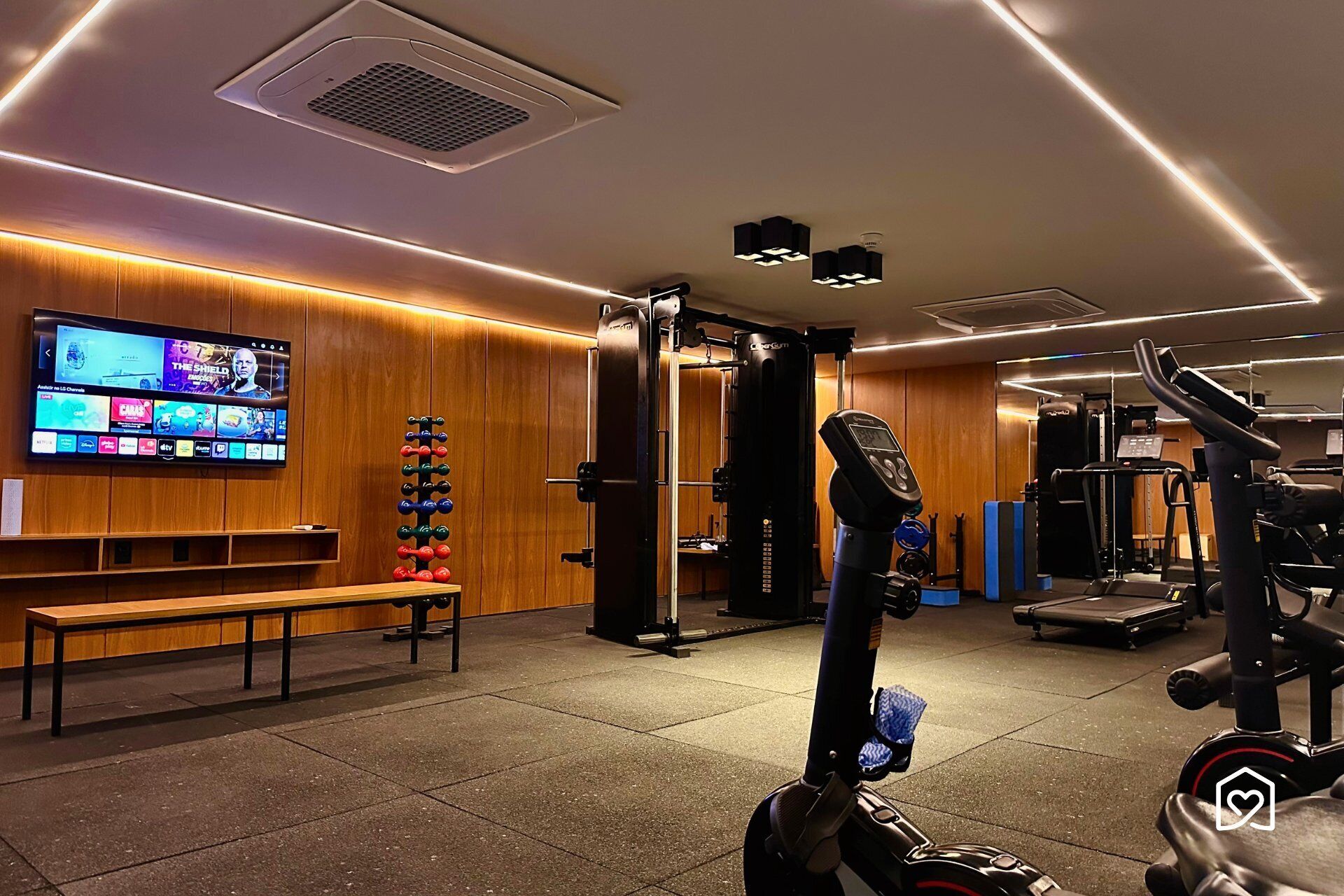 A gym with exercise equipment, a bench, and a TV displaying various channels.