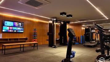A gym with exercise equipment, a bench, and a TV displaying various channels.