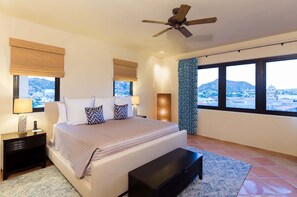 A bedroom with a bed, bedside tables, a nightstand, a ceiling fan, and a view of a cityscape through large windows. - 4BR Medano Beach Penthouse | Pools, Beach Club & Walk to Town (Cabo San Lucas)