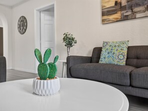 Ein Wohnzimmer mit einem weißen runden Tisch, einem grauen Sofa, einem gemusterten Kissen, einer Kakteenpflanze in einem weißen Topf und einer Wanduhr.