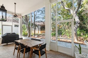 Une terrasse extérieure avec une table ronde et des chaises, un barbecue et une vue sur un jardin arboré agrémenté d’une pergola.