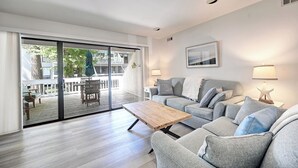 Uma sala de estar moderna com um sofá cinzento, uma mesa de centro de madeira e uma porta de vidro que dá para um deck exterior.
