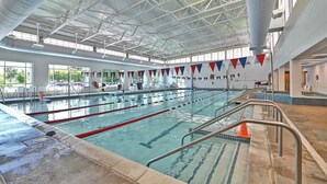 Piscina interna com várias pistas de natação, teto alto com vigas de metal e vista para o exterior através de grandes janelas.