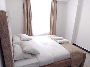 Ein Bett mit weißer Bettwäsche, einem Kopfteil, Vorhängen und einem Fenster mit Tageslicht.