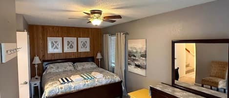 Un dormitorio con cama, una mesita, una silla, un ventilador de techo y un espejo.